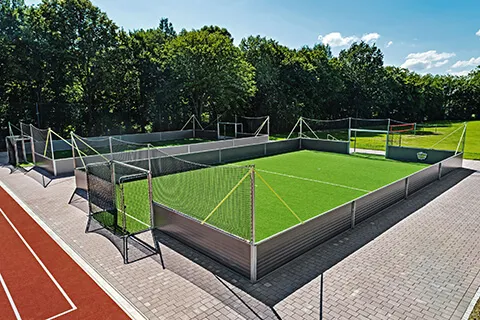 Kleinfeld-Fußballplatz mit grünem Kunstrasen und Umzäunung in einer städtischen Parkumgebung bei klarem Himmel.